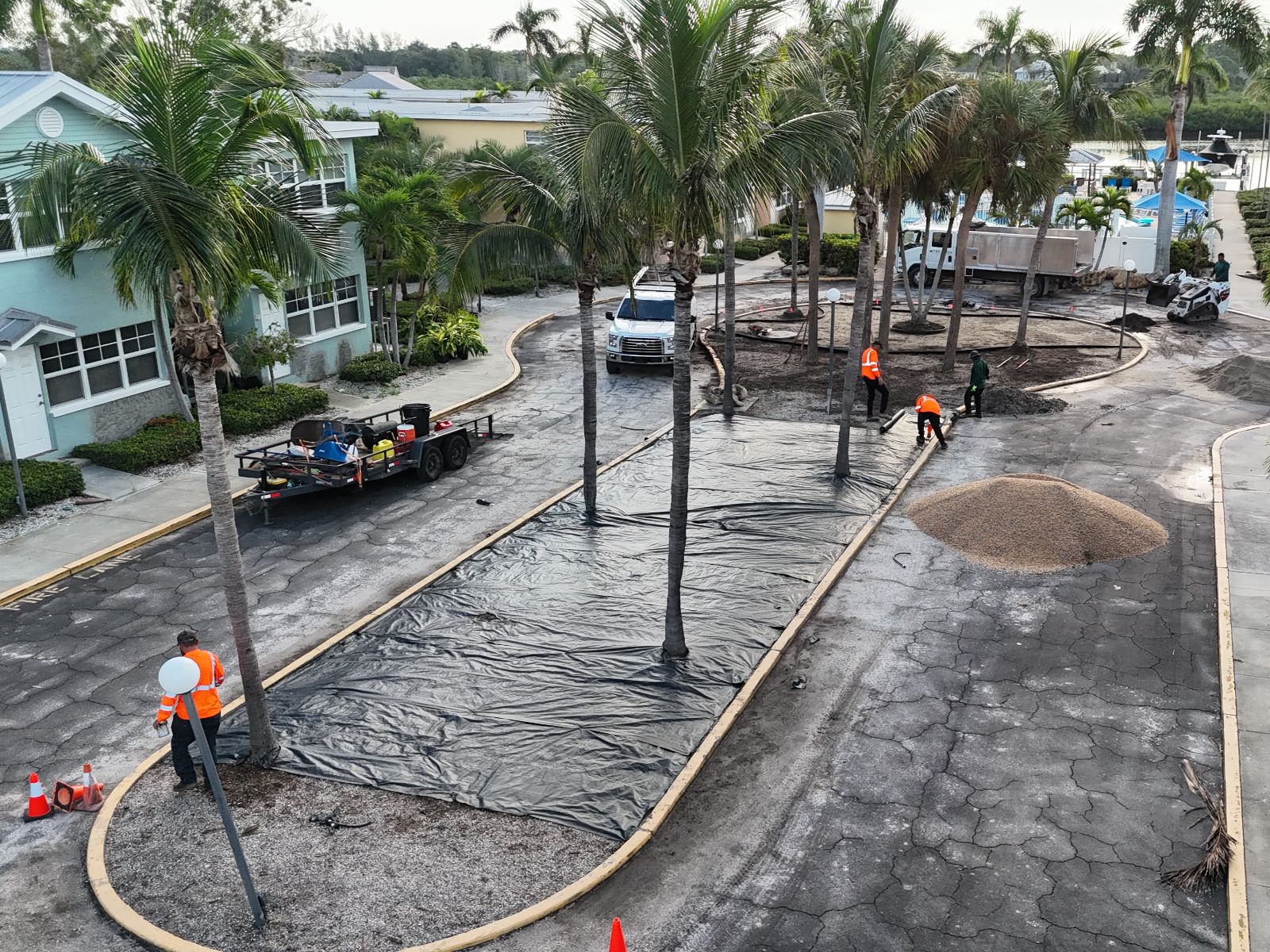Construction workers are landscaping a street, laying down black fabric and gravel around palm trees in a tropical setting.
