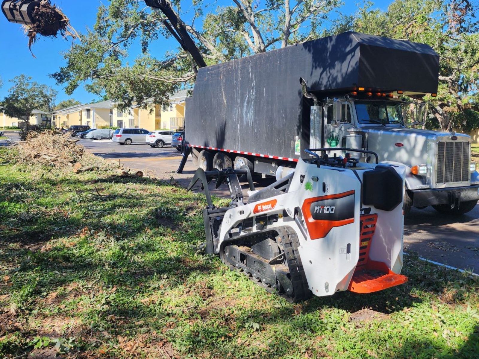 A compact loader and a large truck are parked near tree debris, with buildings and cars visible in the background.