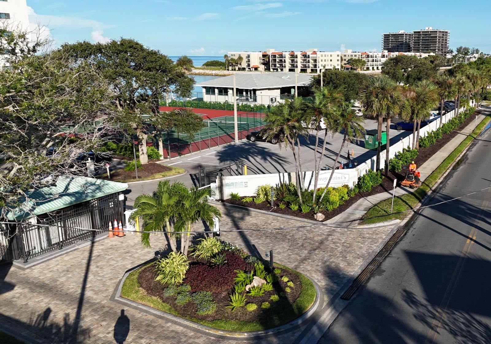 A sunny waterfront area with landscaped gardens, tennis courts, buildings, and maintenance workers on a paved road. Blue sky above.