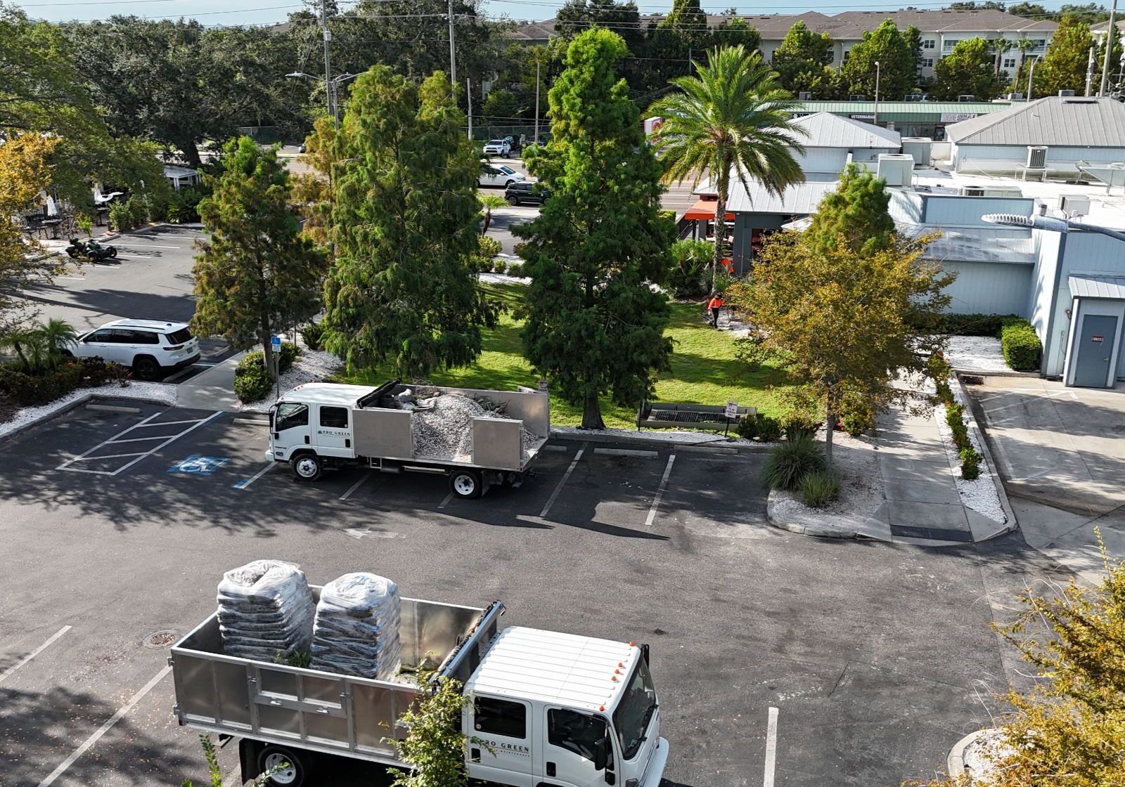 Two trucks loaded with materials parked in a lot, surrounded by trees and buildings in a sunny neighborhood setting.